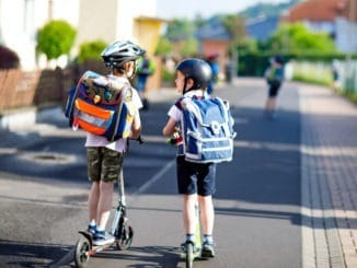 Two school kid boys in safety helmet riding with scooter in the city with backpack on sunny day. Happy children in colorful clothes biking on way to school.