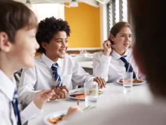 Group Of High School Students Wearing Uniform Sitting Around Table And Eating Lunch In Cafeteria