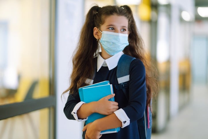 Schoolgirl in a protective mask with a backpack and a textbook in her hands.