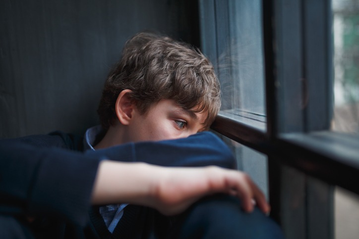 Pensive sad boy teenager in a blue shirt and jeans sitting at the window and closes his face with his hands.