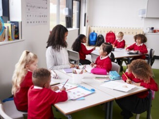 Young female infant school teacher sitting at a table in a classroom with her pupils