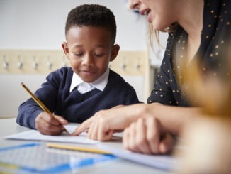 Female primary school teacher helping a young school boy sitting at table in a classroom, close up, selective focus