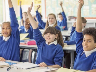 Elementary school children wearing blue school uniforms raising hands in classroom
