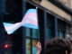 A hand holds up a small transgender pride flag in a Bristol Street. The transgender pride flag is a symbol of pride for the trans community. The blue and pink stripes represent traditional colours for a boy and girl, while the white stripe represents transitioning, intersex, neutral and undefined gender.