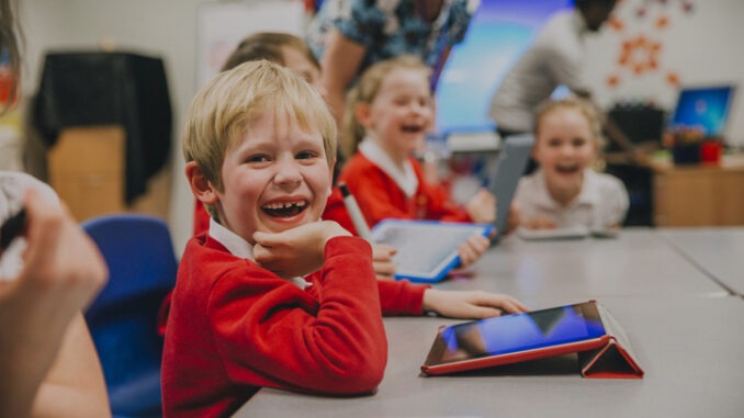 Happy little boy is smiling for the camera while using a digital tablet in his technology lesson at school.