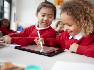 close up of two kindergarten schoolgirls wearing school uniforms, sitting at a desk in a classroom using a tablet computer and stylus, looking at screen and smiling