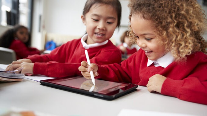 close up of two kindergarten schoolgirls wearing school uniforms, sitting at a desk in a classroom using a tablet computer and stylus, looking at screen and smiling