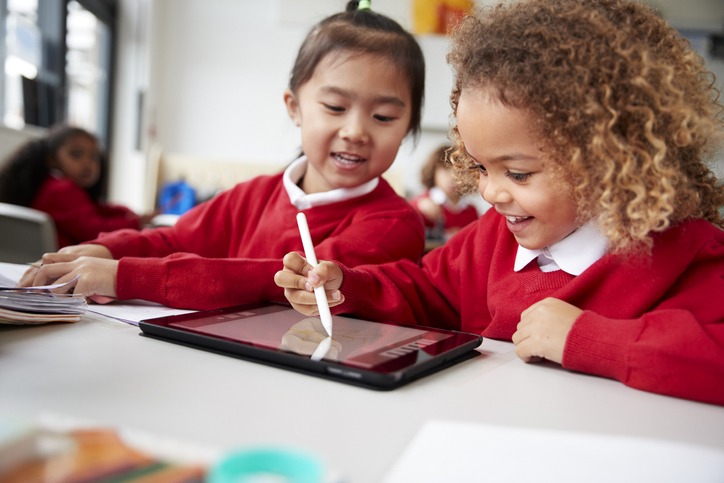 close up of two kindergarten schoolgirls wearing school uniforms, sitting at a desk in a classroom using a tablet computer and stylus, looking at screen and smiling