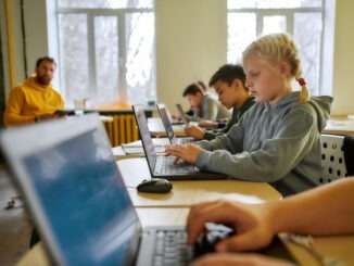 Portrait of caucasian schoolgirl looking at the screen of the laptop together with other pupils during a lesson in modern smart school. Male teacher in the background. STEM disciplines