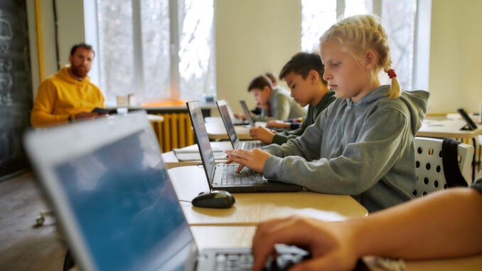 Portrait of caucasian schoolgirl looking at the screen of the laptop together with other pupils during a lesson in modern smart school. Male teacher in the background. STEM disciplines