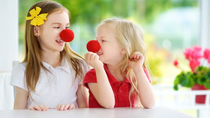 Happy little sisters wearing red clown noses having fun together on sunny summer day at home