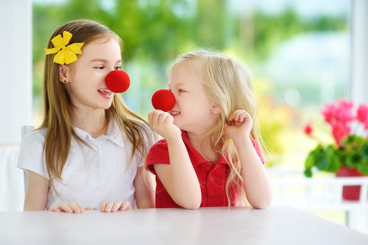 Happy little sisters wearing red clown noses having fun together on sunny summer day at home