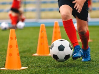 Footballer dribbling ball on training between orange cones. Young football player in sports blue cleats and red socks