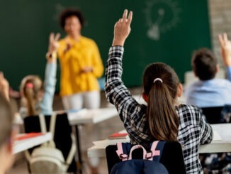Rear view of schoolgirl raising her arm to answer the question in the classroom.