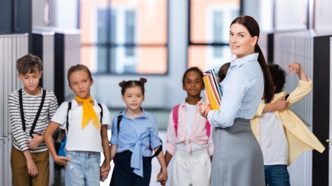 selective focus of teacher looking at camera while standing with multiethnic pupils in school corridor