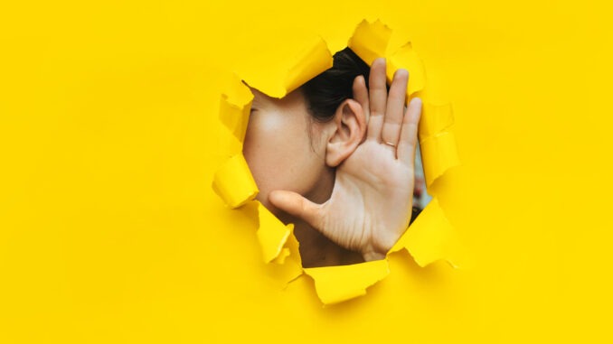 Close-up of a woman's ear and hand through a torn hole in the paper. Yellow background, copy space. The concept of eavesdropping, espionage, gossip and tabloids.