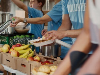 At food drive, poor female wheelchair user receives free food and provisions. Multiethnic volunteers in blue t-shirt distributing fresh fruits and hot meals to less privileged. Close-up, tripod.