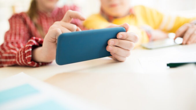 Two modern schoolmates playing in smartphone over desk during break between lessons