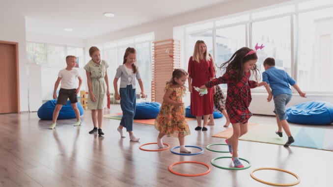 Small nursery school children with female teacher on floor indoors in classroom, doing exercise. Jumping over hula hoop circles track on the floor.