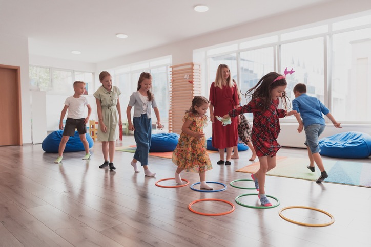 Small nursery school children with female teacher on floor indoors in classroom, doing exercise. Jumping over hula hoop circles track on the floor.