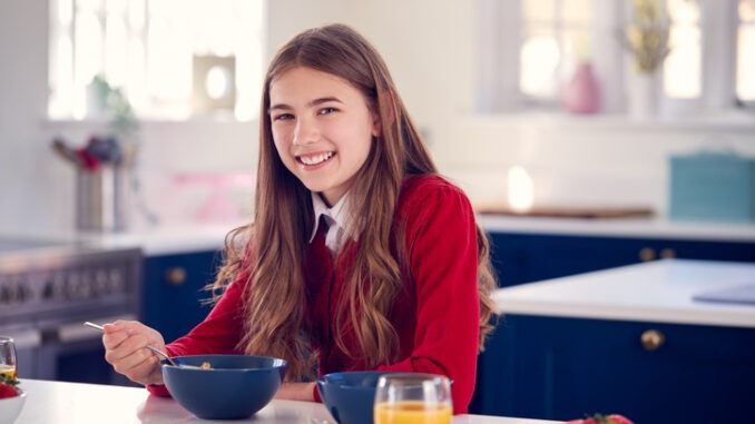 Portrait Of Smiling Teenage Girl Wearing School Uniform In Kitchen Eating Healthy Breakfast