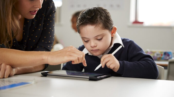 Young female teacher working with a Down syndrome schoolboy sitting at desk using a tablet computer in a primary school classroom, front view, close up