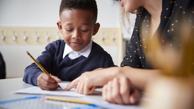Female primary school teacher helping a young school boy sitting at table in a classroom, close up, selective focus