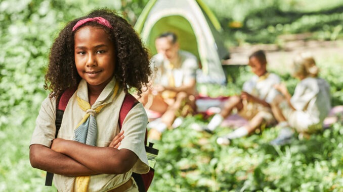 Waist up portrait of cute black girl scout looking at camera while camping with school group