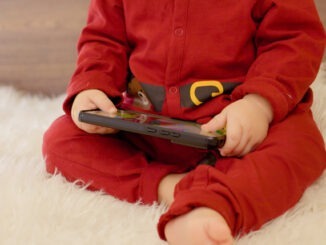 A young boy sits on a soft surface, completely focused on his smartphone. The festive atmosphere is enhanced by a beautifully decorated Christmas tree and colorful ornaments.