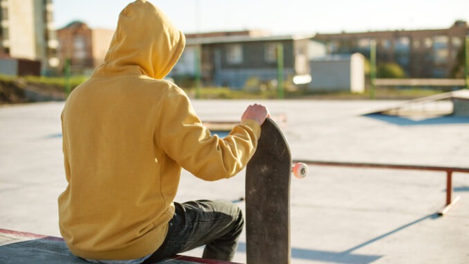Close-up of a young hipster dressed in a sweatshirt and jeans sitting sad in a skate park and holding a skateboard
