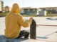 Close-up of a young hipster dressed in a sweatshirt and jeans sitting sad in a skate park and holding a skateboard