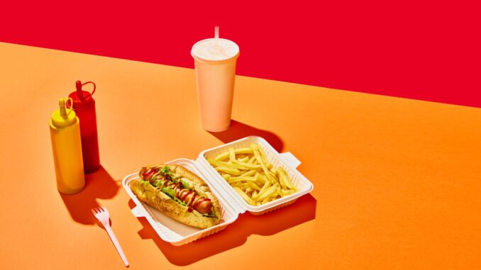 Tasty hotdog with sausage served mustard and ketchup with green salad with refreshing drinks and fried potato against vivid red background. Concept of fast food, street food, delivery. Copy space. Ad