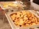 A large tray holds a mound of crispy chicken nuggets, freshly prepared, while another tray nearby features golden fries. This scene is set in a busy cafeteria during lunch.
